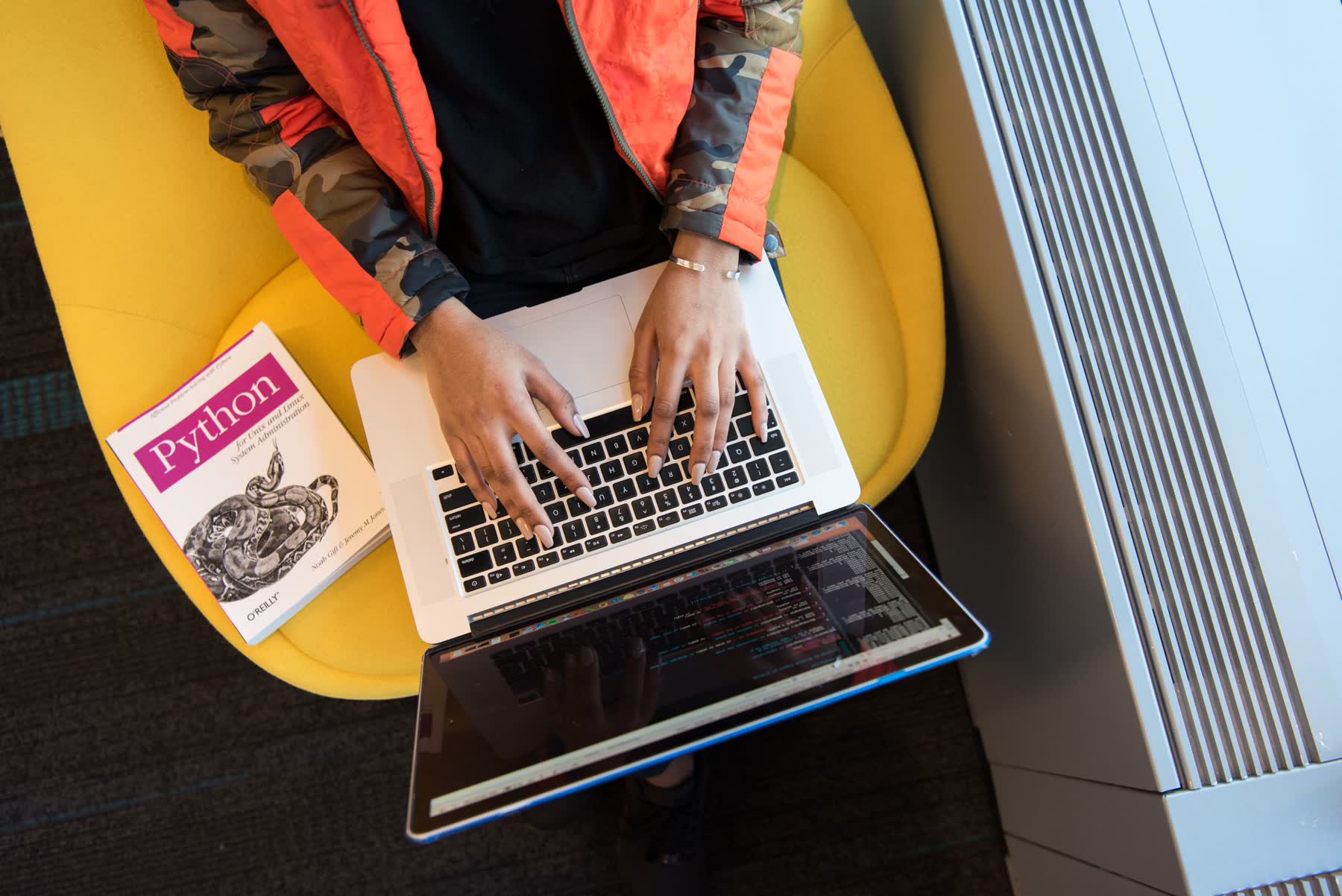 Mulher programando num laptop, com um livro de Python ao lado